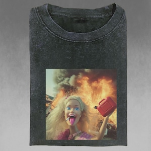 Puede incluir: Camiseta gris oscuro con un gráfico de una muñeca rubia con un cigarrillo y una lata de gasolina roja, sobre un fondo de llamas y humo. La camiseta tiene cuello redondo.