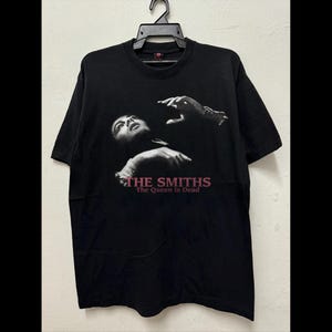 May include: Black t-shirt featuring a grayscale image of a person with their head tilted back and arms raised. The text "THE SMITHS" and "The Queen Is Dead" are printed in pink below the image. The shirt is hanging on a black hanger.