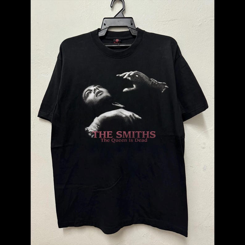 May include: Black t-shirt featuring a grayscale image of a person with their head tilted back and arms raised. The text "THE SMITHS" and "The Queen Is Dead" are printed in pink below the image. The shirt is hanging on a black hanger.