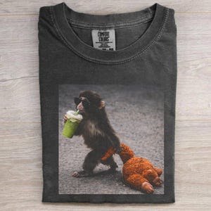 Puede incluir: Camiseta Comfort Colors gris oscuro con un mono con gafas de sol bebiendo una bebida verde con crema batida. El mono sostiene un pequeño peluche naranja. La camiseta tiene cuello redondo y una etiqueta Comfort Colors.