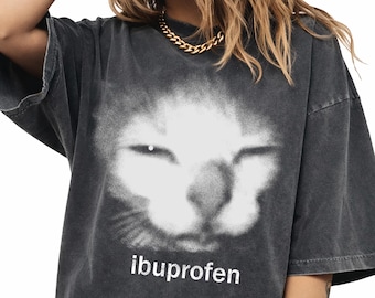May include: Dark gray t-shirt featuring a white, blurred cat face graphic. The word "ibuprofen" is printed in white below the graphic. The shirt has a vintage, washed appearance.