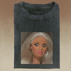 Puede incluir: Camiseta gris oscuro con la cara de una muñeca con expresión de sorpresa. La muñeca tiene pelo largo y blanco, ojos grandes y la boca ligeramente fruncida. La camiseta tiene cuello redondo y un aspecto ligeramente descolorido.