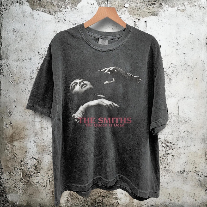 May include: Dark gray t-shirt featuring a black and white image of a person looking up with hands raised. The text "THE SMITHS" and "The Queen Is Dead" are printed in a burgundy color.