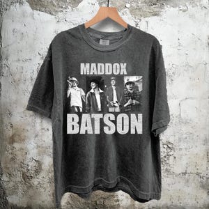 Puede incluir: Camiseta gris oscuro con las palabras blancas "MADDOX BATSON". La camiseta presenta una foto en blanco y negro de cuatro personas. Cuelga de una percha de madera contra una pared texturizada.