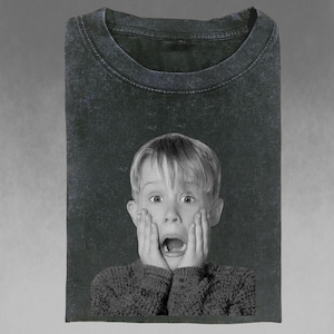 May include: A black and white graphic tee featuring a surprised child with hands on his cheeks and mouth open. The shirt is folded, showing the neckline and a portion of the front graphic. The tee is a dark, faded color.