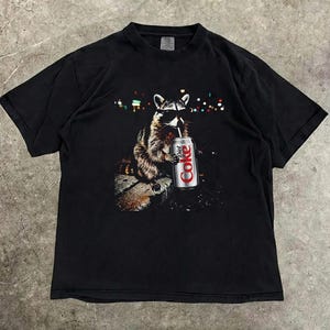 May include: Black t-shirt featuring a raccoon wearing sunglasses and drinking from a Diet Coke can. The raccoon is depicted in a detailed illustration with a blurred background of city lights. The shirt is a classic crew neck.