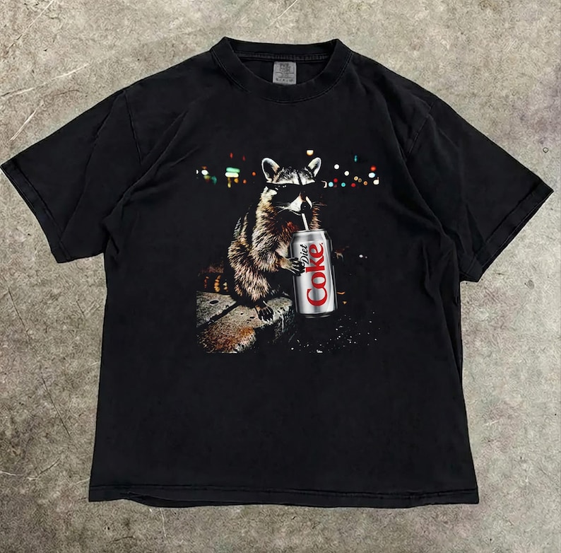 May include: Black t-shirt featuring a raccoon wearing sunglasses and drinking from a Diet Coke can. The raccoon is depicted in a detailed illustration with a blurred background of city lights. The shirt is a classic crew neck.