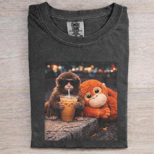 以下が含まれることがあります： 2匹の猿のグラフィックが特徴の濃いグレーのComfort Colors Tシャツ。一匹はサングラスをかけ、アイスコーヒーを飲んでいます。もう一匹はオレンジ色で抱きしめたくなるような猿です。Tシャツは折りたたまれています。