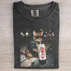 May include: Dark gray t-shirt featuring a cool cat wearing sunglasses, a black leather jacket, and a gold chain, holding a Diet Coke can with a straw. The background shows city lights. The shirt has a Comfort Colors tag.
