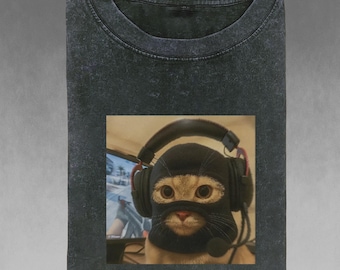 Può includere: T-shirt grigio scuro con un'immagine quadrata di un gatto con passamontagna nero e cuffie da gioco. Il gatto guarda avanti con gli occhi spalancati. Lo sfondo mostra uno schermo di computer e una scrivania.