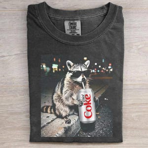 May include: Dark gray Comfort Colors t-shirt featuring a raccoon wearing sunglasses and drinking from a Diet Coke can. The raccoon is set against a blurred city night scene. The Diet Coke can is silver with red text.