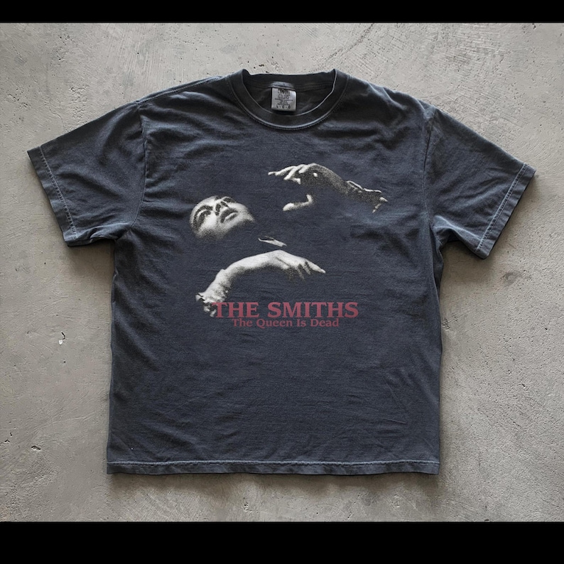 May include: Dark gray t-shirt featuring a black and white image of a person with outstretched hands. The text "THE SMITHS" and "The Queen Is Dead" are printed in red below the image. The shirt has a crew neck and short sleeves.