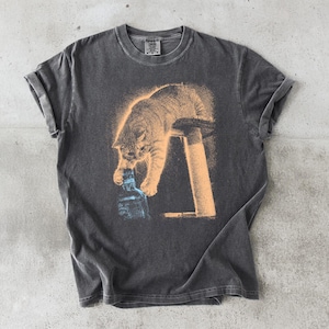 Könnte beinhalten: Ein anthrazitfarbenes Kurzarm-T-Shirt mit einem Grafikdruck einer hellorangen Katze, die auf einem Pfosten sitzt und mit einer Pfote nach einer blauen Glasflasche greift. Das T-Shirt hat einen Rundhalsausschnitt und eine Vintage-Waschung.