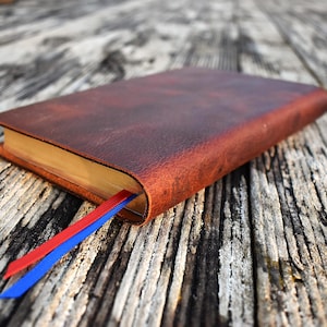 May include: A brown leather-bound journal with a red and blue ribbon bookmark. The journal's pages have a golden hue, and the cover shows a textured surface. The journal rests on a weathered wooden surface.