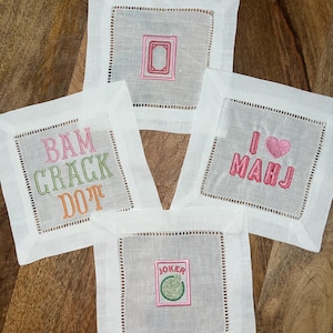 以下が含まれることがあります： 刺繍デザインが施された4つの白いリネンコースター。1つはピンクのフレーム、もう1つは緑、ピンク、オレンジで「BAM CRACK DOT」と書かれています。3つ目はピンクで「I HEART MAHJ」、最後は「JOKER」と書かれています。