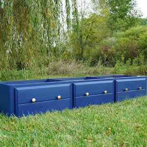 May include: A navy blue wooden storage unit with three sections, each featuring a drawer with a gold-coloured knob. The unit is placed on a grassy lawn, with a backdrop of trees and foliage.