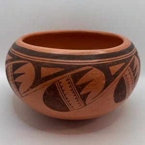 May include: A round terracotta clay bowl featuring a wide, open top and a smooth, matte finish. The exterior is decorated with dark brown, hand-painted geometric patterns, including repeating leaf-like shapes and thin, vertical dashed lines.