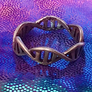 May include: A silver-toned ring with a DNA double helix design. The ring is made of metal and features a repeating pattern of vertical bars and curved lines. The ring is displayed on a shiny, iridescent purple and blue surface.