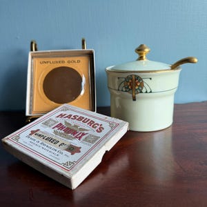 May include: A cream-colored ceramic lidded jar with a gold-toned handle and spoon, alongside a square box labeled Hasburg's Phoenix Unfluxed Gold and a matching circular gold disc in a display frame, featuring decorative geometric floral motifs.