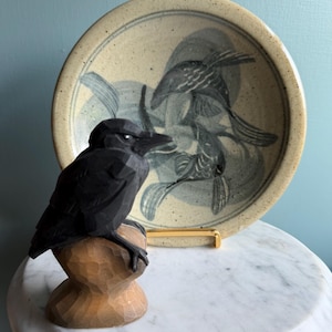 May include: A hand-carved black wooden crow figurine perched on a natural wood stump, positioned in front of a speckled beige ceramic plate featuring a dark blue painted design of two fish swimming in a circular motion on a gold metal stand.