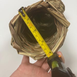 May include: A cylindrical woven straw container with a hollow center. A yellow measuring tape is placed across the opening, showing a diameter of approximately 4 inches. The natural tan fibers are woven in a crisscross pattern throughout.