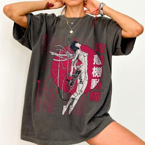May include: Dark gray t-shirt featuring a graphic of a cyborg woman with a red circle and Japanese text. The design includes red lines and a futuristic aesthetic. The shirt has a relaxed fit.