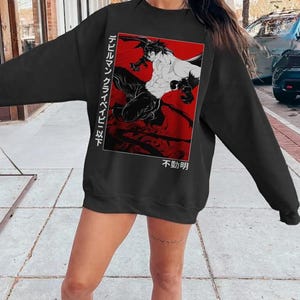 May include: Black sweatshirt featuring a red and white graphic of a manga-style illustration. The graphic depicts two figures in action, with Japanese text on the left and bottom right. The sweatshirt is a long-sleeved crew neck.