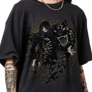 May include: Dark gray t-shirt featuring a detailed graphic of a character with long hair and multiple eyes, accompanied by a wolf-like creature with many eyes. The design is in black and gold tones, creating a striking visual.