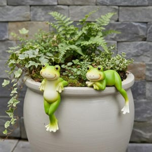 Puede incluir: Una maceta grande de cerámica gris claro llena de vegetación, con dos figuras de ranas verdes. Una rana está de pie, la otra descansa en el borde de la maceta. Las ranas tienen patas blancas y caras sonrientes. El fondo es una pared de piedra gris.