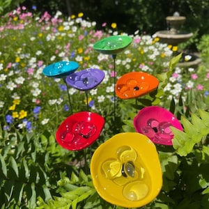 May include: A selection of colourful glass flower garden stakes. Each flower has a vibrant hue, including green, blue, purple, orange, red, pink, and yellow, with clear glass marbles in the centre. The stakes are set in a lush garden.