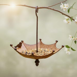 May include: A bronze-colored, umbrella-shaped bird feeder with a small bird perched on the side. The feeder is filled with birdseed and hangs from a curved metal hook. The feeder is suspended from a tree branch with white flowers.