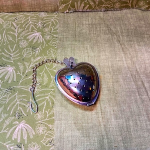 May include: A heart-shaped, silver tea infuser with a chain and hook. The infuser has small holes and is resting on a green fabric background. The background has a floral pattern on the left side.