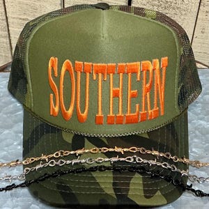 May include: A green camouflage trucker hat with the word "SOUTHERN" embroidered in orange. The hat has a mesh back and a curved brim. Several strands of barbed wire necklaces are in the foreground.