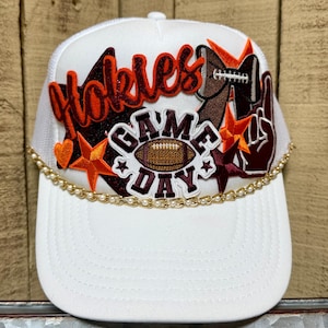 May include: White trucker hat with a curved brim, featuring the word "Hokies" in orange and brown, along with "Game Day" and football-themed embellishments. A gold chain with pearls accents the brim.