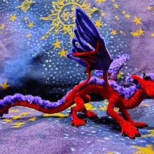 May include: A handmade dragon figurine crafted from red and purple pipe cleaners. The dragon has large wings, a long tail, and is posed on a starry background. The dragon's wings and back are purple, while the body is red.