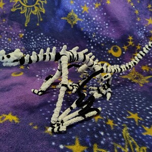 May include: A handmade dinosaur skeleton sculpture crafted from white and black pipe cleaners. The dinosaur is posed on a purple and blue celestial patterned fabric with gold stars and suns.