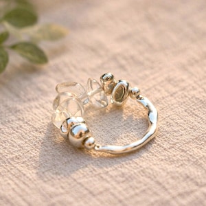 May include: A silver ring with clear, irregular-shaped stones and small silver beads. The ring has a textured band and is set against a neutral background. The ring is a piece of jewelry.