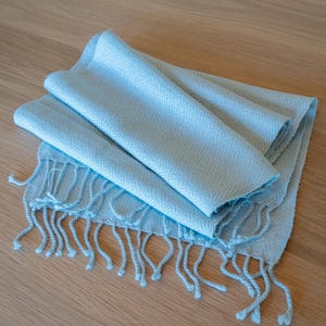 May include: A folded, light blue scarf with a fringed edge. The scarf is made of a soft, woven material and is lying on a light brown wooden surface. The scarf is a solid color and has a simple, elegant design.