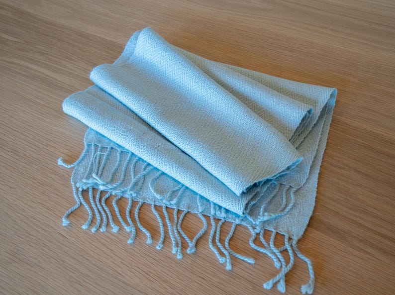 May include: A folded, light blue scarf with a fringed edge. The scarf is made of a soft, woven material and is lying on a light brown wooden surface. The scarf is a solid color and has a simple, elegant design.