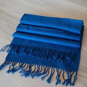 May include: A folded, blue scarf with a fringed edge. The scarf is a solid, deep blue color and appears to be made of a soft material. The fringe is the same blue color as the scarf. The scarf is lying on a light-colored wooden surface.
