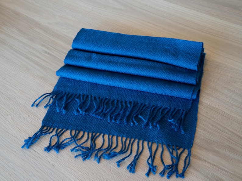 May include: A folded, blue scarf with a fringed edge. The scarf is a solid, deep blue color and appears to be made of a soft material. The fringe is the same blue color as the scarf. The scarf is lying on a light-colored wooden surface.