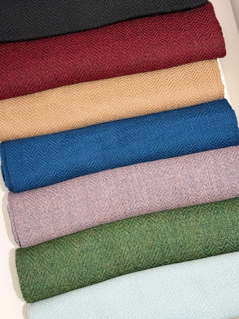 May include: A collection of folded fabric rolls in various colors, including black, burgundy, tan, blue, lavender, green, and light blue. Each roll displays a textured herringbone pattern, suggesting a woven textile material.