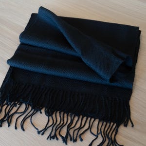 May include: A dark navy blue scarf with a herringbone pattern and fringe. The scarf is folded and draped, showcasing its texture and design. The fringe hangs loosely at the bottom.