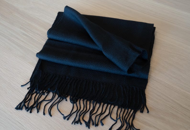 May include: A dark navy blue scarf with a herringbone pattern and fringe. The scarf is folded and draped, showcasing its texture and design. The fringe hangs loosely at the bottom.