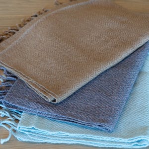 May include: A stack of three woven scarves in shades of tan, blue-grey, and light blue. Each scarf has a herringbone pattern and fringed edges. The scarves are neatly folded and layered on a light wood surface.