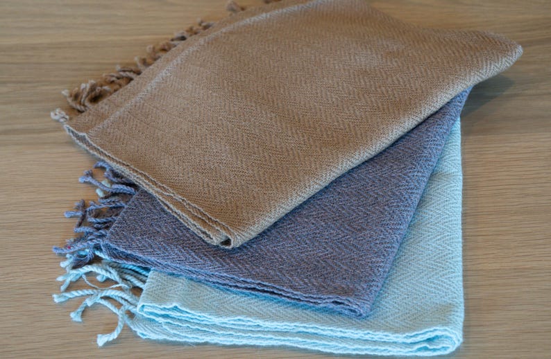 May include: A stack of three woven scarves in shades of tan, blue-grey, and light blue. Each scarf has a herringbone pattern and fringed edges. The scarves are neatly folded and layered on a light wood surface.