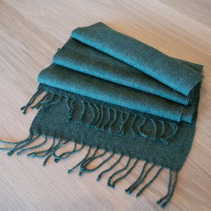 May include: Folded teal scarf with fringe detail. The scarf is made of a soft, woven material. The scarf is folded in thirds, with the fringe at the bottom. The scarf is lying on a light wood surface.