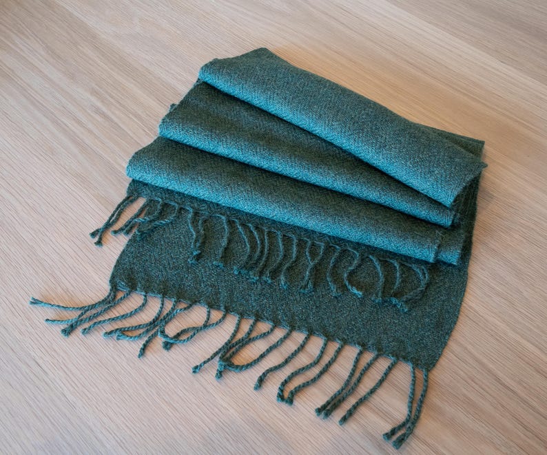 May include: Folded teal scarf with fringe detail. The scarf is made of a soft, woven material. The scarf is folded in thirds, with the fringe at the bottom. The scarf is lying on a light wood surface.
