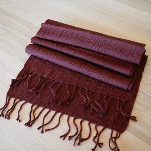 May include: A folded burgundy scarf with a herringbone pattern and fringe. The scarf is made of a soft material and is displayed on a light wood surface. The scarf is a deep red color.