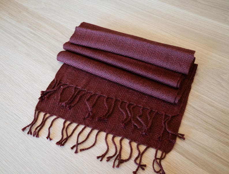 May include: A folded burgundy scarf with a herringbone pattern and fringe. The scarf is made of a soft material and is displayed on a light wood surface. The scarf is a deep red color.